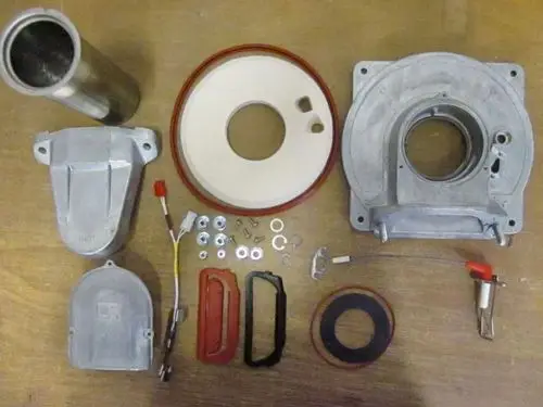 Zerlegte Teile eines Wärmetauschers von Remeha, darunter Aluminiumdeckel, Dichtungen, Metallrohre, Schrauben und elektrische Komponenten auf einem Holztisch.